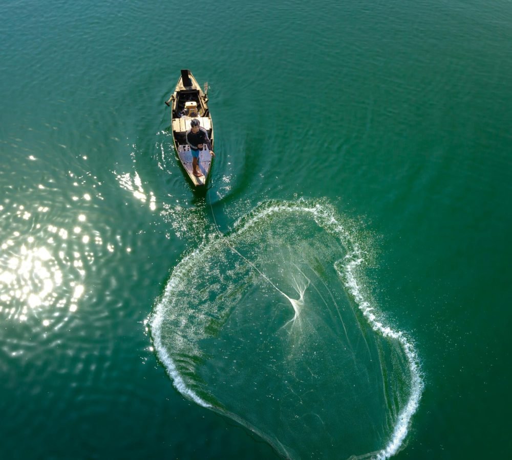 pexels-photo-2150403-2150403 Aerial shot of a fisherman casting a net from a boat in serene, sunlit waters.